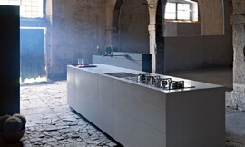 Kubistisch gevormd keukenblok. Platte, greeploze fronten met subtiel aanrechtblad in dezelfde grijstint. Keukenkraan komt uit een achterwand. Gootsteen afdekbaar. Keuken op sokkel. Zuordnung: Stil Design-keukens, Planungsart keukenblok