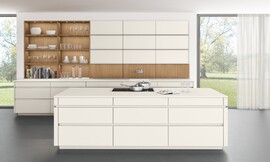 Een witte keuken met greeploze matte lakfronten die er vrijwel niet meer als keuken uitziet, doordat de apparatuur nauwelijks zichtbaar is. Dit maakt deze keuken met een hoog keukenblok aan de wand en een apart kookeiland uitermate geschikt om als open keuken naadloos aan te sluiten op de woonruimte. Ook de afwisseling tussen het tijdloze wit en het koperkleurige eiken uit het programma TIMBER zorgt voor een sfeervol en warm woonaccent. Zuordnung: Stil Moderne keukens, Planungsart L-vormige keuken