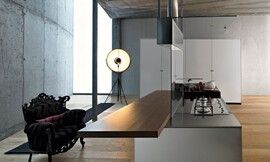 Design kookeiland met wit glanzende fronten (greeploos). Een design afzuigkap is verbonden met het kookeiland middels een glasplaat. Aan de achterkant zit een houten aanrechtblad dat tegelijkertijd als bar/zitgelegenheid dienst doet. Dunne wangen en aanrechtblad. Zuordnung: Stil Moderne keukens, Planungsart Keuken met keukeneiland