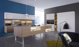 De combinatie van bijzonder natuurlijk werkende kopereiken fronten met matte sahara-lak zorgt voor een interessante spanning. In de hangkast is een efficiënte afzuigkap geïntegreerd die geen luchtafvoer behoeft. De keuken bestaat uit drie delen: het opgehangen keukenblok met fornuis en spoelbak, de eikenhouten fronten en daarboven de witte hangkasten; het keukeneiland in het midden, staande op metalen open poten die het geheel daardoor een luchtig karakter geven en de hoge witte kasten aan de andere wand waarin op praktische werkhoogte de roestvrijstalen apparatuur is aangebracht. Zuordnung: Stil Moderne keukens, Planungsart L-vormige keuken