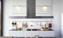 Designkeuken met symmetrische witte paneelwand waar overzichtelijk handige dingen als een kookboekstandaard, kruidenpotjes, messenrek of keukenpapierhouder aan kunnen worden gehangen. Bij deze keuken loopt de wand door tot aan het plafond. Vrij hoog zijn er nog twee spotjes geplaatst, maar ook op de hoogte van de kookboekhouder etc. zijn er kleine spotjes opgenomen. Dit zorgt voor een goede verlichting op het aanrechtblad en de dubbele spoelbak daaronder. De onderkastjes zijn greeploos en wit. Het aanrechtblad is dun en heeft een donkere kleur. Het kookeiland daar tegenover biedt ook naast het gasfornuis genoeg plaats voor het snijden, hakken en mengen van interessante recepten. Zuordnung: Stil Design-keukens, Planungsart U-vormige keuken