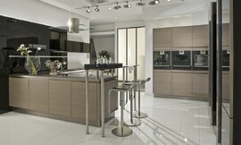 De apparatuur is in deze moderne keuken met greeploze eiken houten fronten op praktische werkhoogte in hoge kasten aan de wand opgenomen. Levendig en modern wordt de keuken door het wandpaneel dat achter het L-vormige keukenblok is bevestigd: dit is namelijk hoogglanzend zwart en biedt de mogelijkheid legplanken op te nemen. Het L-vormige gedeelte van de keuken bestaat allereerst uit een blok dat aan de wand is geplaatst, met spoelbak en daarboven lage hoogglanzende zwarte bovenkasten. Om de hoek is dan het kookeiland waarop het fornuis is bevestigd, daarboven hangt de moderne afzuigkap van roestvrij staal en glas. Aan het eind van het kookeiland is een bar opgenomen die aan de ene kant op twee korte, ronde metalen poten op het aanrechtblad rust en aan de andere kant op twee hoge poten staat. De zitgelegenheid die hierdoor is ontstaan biedt plaats aan twee personen. Zuordnung: Stil Landelijke keukens, Planungsart U-vormige keuken