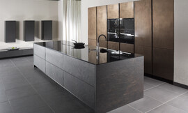 De open ontworpen, representatieve luxe keuken biedt maximale functionaliteit met veel bergruimte. Voor het kookeiliand is gebruik gemaakt van echte grijze leisteen. Het dunne, hoogglanzende aanrechtblad zorgt voor een interessant contrast. Op het kookeiland zijn de spoelbak en het fornuis opgenomen. In de wand zijn in hoge kasten de apparaten opgenomen, twee daarvan op een praktische werkhoogte. De luxe en exclusieve uitstraling van deze greeploze keuken wordt door niets verstoord. Zuordnung: Stil Luxe keukens, Planungsart Detail keukenontwerp