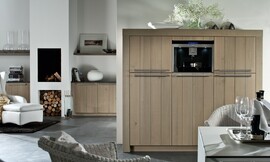 Hier ziet u een detailfoto van de hoogkast/apparatuurkast met brede wangen, stanggrepen en verticale strepen in het hout. Ook ziet u hoe hetzelfde concept is doorgevoerd in de woonkamer. Aparte eettafel met rieten stoelen. Zuordnung: Stil Landelijke keukens, Planungsart L-vormige keuken