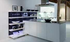 Deze open keuken heeft een gereduceerde look met witte fronten en een dun, metallic aanrechtblad. Het kookeiland staat haaks op een open wandgedeelte. Deze open wand met legplanken is ideaal om alles direct bij de hand te hebben, maar kan tegelijkertijd ook decoratief worden gebruikt. De blauwe achtergrond van de open kast zorgt voor een speels accent. Op deze open wandkast sluit het keukenblok met de spoelbak aan. De onder- en bovenkasten bieden voldoende bergruimte. Een keukenplanning zoals deze is ook erg geschikt voor grote ruimtes die deels als woonkamer functioneren. Zuordnung: Stil Design-keukens, Planungsart Open keuken (woonkeuken)