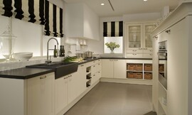 Landelijke L-vormige keuken met moderne touch. Hier zijn de witte cassettefronten gecombineerd met een contrasterend zwart aanrechtblad. De spoelbak heeft dezelfde kleur, maar is iets hoger dan het aanrechtblad geplaatst. In hetzelfde keukenblok als de spoelbak is ook het fornuis opgenomen. In dit deel van de keuken zijn er alleen onderkasten, voor een deel met praktische grote lades, een ander deel met deurtjes en er is ook een praktisch wijnrek beschikbaar voor de wijnliefhebber. Om de hoek is er eerst een onderkast met twee deurtjes, daarna twee open onderkasten met manden, die het geheel een landelijke sfeer geven. Op het aanrechtblad daar boven zijn twee hoge kasten met fraaie sierlijsten gezet. De glazen deurtjes van de twee kasten maken het mogelijk een blik te werpen op het mooie servies, de bijzondere wijnglazen of andere interessante keukenaccessoires. Onder de kast met glazen deuren zijn er nog vier handige lades aangebracht. Als los element is er een apparatuurkast geplaatst. Ook deze kast wordt aan de bovenkant gesierd door een mooie afsluitende lijst. In het midden van dit keukenelement bevindt zich op handige werkhoogte de oven. Direct daaronder is er een open kast met legplank. Zuordnung: Stil Landelijke keukens, Planungsart L-vormige keuken