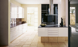 Witte design keuken met hoogglans fronten en een zwarte hoogkast voor apparatuur. De keuken bestaat uit een keukenblok dat tegen de wand is geplaatst en een afzonderlijk kookeiland. De spoelbak bevindt zich in het wandgedeelte. Tevens zijn hier vele kasten geplaatst, zodat er voldoende bergruimte is ontstaan. De onderkasten en lades zijn voorzien van horizontale, grote grepen. De bovenkasten hebben verticaal geplaatste grepen. Het kookeiland heeft evenals het keukenblok met de spoelbak een dun granieten werkblad in een contrasterende donkere kleur. Achter het kookeiland is de apparatuur in hoge, glanzende zwarte kasten opgenomen. De robuuste vloertegels zijn ook in de sokkel van het kookeiland verwerkt, zodat er een eenheid ontstaat. Zuordnung: Stil Design-keukens, Planungsart Keuken met zitgelegenheid