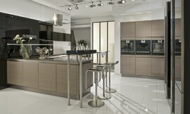 De apparatuur is in deze moderne keuken met greeploze eiken houten fronten op praktische werkhoogte in hoge kasten aan de wand opgenomen. Levendig en modern wordt de keuken door het wandpaneel dat achter het L-vormige keukenblok is bevestigd: dit is namelijk hoogglanzend zwart en biedt de mogelijkheid legplanken op te nemen. Het L-vormige gedeelte van de keuken bestaat allereerst uit een blok dat aan de wand is geplaatst, met spoelbak en daarboven lage hoogglanzende zwarte bovenkasten. Om de hoek is dan het kookeiland waarop het fornuis is bevestigd, daarboven hangt de moderne afzuigkap van roestvrij staal en glas. Aan het eind van het kookeiland is een bar opgenomen die aan de ene kant op twee korte, ronde metalen poten op het aanrechtblad rust en aan de andere kant op twee hoge poten staat. De zitgelegenheid die hierdoor is ontstaan biedt plaats aan twee personen. Zuordnung: Stil Moderne keukens, Planungsart Keuken met zitgelegenheid