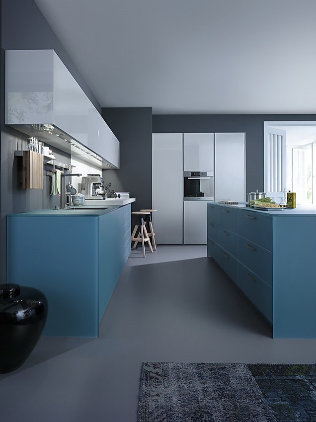 Deze uit drie blokken bestaande keuken met kookeiland bestaat uit mat glas en greeploze fronten met hoogglans lak. De kasten van het kookeiland, vijf stuk in totaal, werken kwadratisch in fris gedempt blauw, de kleur die ook doorloopt in het werkblad. Ook de kasten die daar tegenover staan zetten de frisse blauwe tint voort. Daarboven hangen langwerpige, maar lage kasten met een lichtgrijze hoogglans lak. Haaks op de beide keukenblokken staat het lichtgrijze keukenelement met twee hoge kasten met daartussen een onderkast, de roestvrijstalen oven en een bovenkast. Mat glas (gedempt blauw) en hoogglans lak (lichtgrijs) zijn de homogene tinten van deze designkeuken. Zuordnung: Stil Moderne keukens, Planungsart U-vormige keuken
