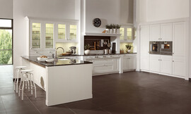 Deze witte keuken laat de moderne interpretatie van een landelijke keuken zien. Heldere lijnvoering, lichte tinten en verfijnde houtoppervlakten. De keuken bestaat uit drie delen: Tegen de wand zijn hoge kasten geplaatst waarin op praktische werkhoogte de roestvrijstalen apparatuur is opgenomen. Tegen de andere wand is een luxe fornuis in een schouw geplaatst, wat bijdraagt aan de landelijke en klassieke uitstraling van de keuken. Naast de schouw zijn aan allebei de kanten twee onderkasten met daarboven twee hangkasten met glazen deurtjes opgenomen. Links sluit een hoge opzetkast aan met dezelfde glazen deuren. Deze opzetkast is geplaatst op het keukeneiland. De spoelbak bevindt zich op het schiereiland. Aan de andere kant van het eiland is door het fraaie zwarte werkblad dat iets uitsteekt een ruime zitgelegenheid ontstaan. Al met al een keuken die aan alle moderne eisen voldoet en desondanks door de schouw en de cassettenfronten een klassieke en landelijke charme biedt. Zuordnung: Stil Landelijke keukens, Planungsart Keuken met keukeneiland