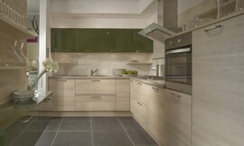 Bovenkasten met olijfgroene hoogglans gelakte fronten. Daaronder licht houten keukenmeubelen met een even licht zandkleurig aanrechtblad. Voor de keukenachterwand tussen keukenblok en bovenkast is geribbeld glas gebruikt. Zuordnung: Stil Moderne keukens, Planungsart keukenblok