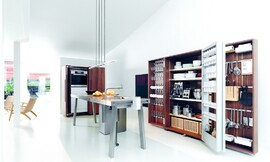 De keukenwerkplaats b2 in actie. De apparaatkast links achter heeft deuren die in de kast ingeklapt kunnen worden waardoor ze niet in de weg zitten tijdens het koken. De opbergkast rechts, daarentegen, heeft omvangrijke opbergmogelijkheden in zijn deuren. Dit concept maakt deze keuken tot een werkplaats. Zuordnung: Stil Design-keukens, Planungsart Keuken met keukeneiland
