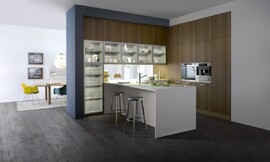 Een harmonieuze drieklank van materialen: keukenfronten met natuurlijk werkend houten structuuroppervlakte, matte magnolia kunststof en een dun wit aanrechtblad/keukenblok. Glazen deurtjes voor de servieskast maken het geheel af. Zuordnung: Stil Klassieke keukens, Planungsart Keuken met keukeneiland
