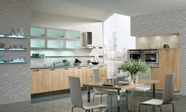 Het lichte hout van de fronten contrasteert met de lichtblauwe glazen bovenkast, de reflecterende vloer en de speelse accenten in het behang. Klassieke keuken met aparte eetkamer- of keukentafel. Zuordnung: Stil Klassieke keukens, Planungsart Keuken met keukeneiland