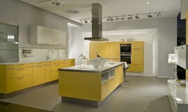 Eilandkeuken met fronten in de opvallende kleur mangogeel. De onderkasten van het keukenblok tegen de wand zijn geel, er zijn zowel lades als kasten. Het spatblad in de nis is wit, net als het aanrechtblad en de twee hangkasten boven de spoelbak. Het tweede keukenelement bestaat uit half hoge kasten waarin ook
de apparatuur is opgenomen. Ook deze kasten zijn allemaal mangogeel. Het kookeiland, eveneens mangogeel, heeft als fris accent ook een wit werkblad. Aan de zijkant steekt het werkbald iets uit waardoor er een zitgelegenheid voor twee personen is ontstaan. Onder het werkblad is er in het kookeiland nog eens extra bergruimte in twee normale kasten, twee lage kastjes en twee open kasten. Zuordnung: Stil Moderne keukens, Planungsart Keuken met keukeneiland