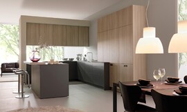 Dit simpele, rechtlijnige ontwerp bereikt een rustige werking van de ruimte. De verschillende tinten (licht hout, donkerbruin, mat zwart) zijn op gewaagde wijze ingezet met een sensibel resultaat: een bijzonder harmonieuze keuken. Een klein aanrechtblad dat over de rand van het keukeneiland uitsteekt creëert een zitgelegenheid. Zuordnung: Stil Moderne keukens, Planungsart U-vormige keuken