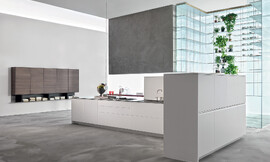 Strakke lijnen zonder onnodige versierselen, helder wit en een moderne uitstraling zijn de kenmerken van deze keuken in eilandvorm. Het rechte keukenblok bestaat uit onderkasten met deurtjes en grote lades die ideaal zijn voor het opbergen van serviesgoed, potten en pannen. De rechterhoek van de keuken wordt omsloten door half hoge kasten. Op de achtergrond is een zeer bijzonder glazen open kastsysteem te zien dat aan het plafond is opgehangen. Zuordnung: Stil Design-keukens, Planungsart Detail keukenontwerp