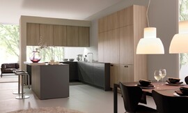 Dit simpele, rechtlijnige ontwerp bereikt een rustige werking van de ruimte. De verschillende tinten (licht hout, donkerbruin, mat zwart) zijn op gewaagde wijze ingezet met een sensibel resultaat: een bijzonder harmonieuze keuken. Een klein aanrechtblad dat over de rand van het keukeneiland uitsteekt creëert een zitgelegenheid. Zuordnung: Stil Moderne keukens, Planungsart U-vormige keuken