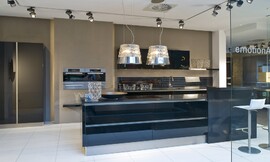 Deze moderne zwarte eilandkeuken in hoogglans uitvoering biedt behalve het kookeiland een keukenblok aan de wand met de spoelbak, onder- en bovenkasten. De bar aan het kookeiland biedt zitgelegenheid voor twee personen. De apparatuur is op praktische werkhoogte in de wand opgenomen. De koel- vriescombinatie is in separaat geplaatste hoge kasten geplaatst. De greeploze zwarte hoogglans fronten reflecteren licht en het patroon van de vloer. Hier ziet u ook een dubbele afzuigkap in een lampenkap. Zuordnung: Stil Moderne keukens, Planungsart keukenblok