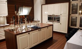 Aanrechtbladen van donker hout en mat witte cassette fronten. Een design afzuigkap geeft het klassieke ontwerp een modern randje. Een symmetrische hoogkast heeft vakken voor twee apparaten. Ook bijzonder is het feit dat houtoptiek voor de volledige wand gebruikt is. Zuordnung: Stil Klassieke keukens, Planungsart Keuken met keukeneiland