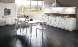 L-keuken in landelijke stijl met cassettefronten. Witte meubels met grijs werkblad. Zitgelegenheid aan vrijstaande tafel. Boven het werkblad is een schouw met ingebouwde afzuigkap aangebracht, die bijdraagt aan het symmetrische karakter van het meubel. Zuordnung: Stil Landelijke keukens, Planungsart L-vormige keuken