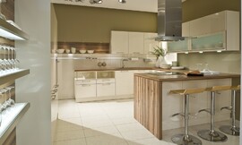 In deze U-vormige keuken is gebruik gemaakt van frisse witte fronten gecombineerd met een houten aanrechtblad dat aan de zijkant van het kookeiland doorloopt en ook als achtergrond bij een legplank is gebruikt. Aan de linkerkant zijn twee onderkasten opgenomen waarvan het bovenste gedeelte open is. Daarnaast komt de spoelkast en nog een onderkast. Boven de spoelbak zijn drie hangkastjes opgenomen. Links van de hangkastjes is een legplank bevestigd tegen hetzelfde hout als het aanrechtblad. Het andere wandgedeelte bestaat uit onderkasten, handige werkruimte op het aanrechtblad met daarboven twee brede kasten met glazen inzet en daaronder spotjes. Haaks hierop staat het kookeiland met fornuis, afzuigkap en aan de andere kant een gezellige zitgelegenheid. In de hoge wandkasten is de apparatuur opgenomen. Zuordnung: Stil Landelijke keukens, Planungsart U-vormige keuken
