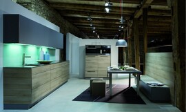 Deze design keuken in L-vorm combineert licht hout (zand) voor de greeploze fronten van de onderkast met donker/paars-blauwe fronten van de bovenkasten. Het aanrechtblad heeft een donkerdere bruintint en loopt als sideboard om de kast heen. Een verlichte glazen plaat geeft de keuken een fonkelend randje. Zuordnung: Stil Moderne keukens, Planungsart keukenblok