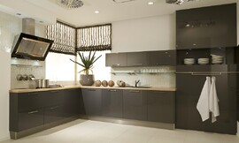 Deze moderne L-vormige keuken heeft donkere antraciet fronten (hoogglans lak) met brede grepen. Deze contrasteren met een zandkleurig aanrechtblad. Het fornuis bevindt zich aan de ene kant van de keuken, met daaronder handige grote lades en de spoelbak bevindt zich aan de andere kant. Boven de spoelbak zijn drie hangkastjes bevestigd. Daaronder is een glazen legplank aan de muur geplaatst. Ideaal om decoratief te gebruiken, of tegelijkertijd handig met mooie glazen. De keuken wordt afgesloten door hoge kasten met op ooghoogte een open gedeelte. De afzuigkap valt door zijn bijzondere design zeker op. Zuordnung: Stil Moderne keukens, Planungsart keukenblok