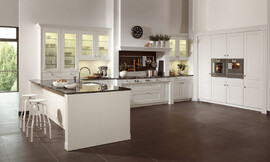 Deze witte keuken laat de moderne interpretatie van een landelijke keuken zien. Heldere lijnvoering, lichte tinten en verfijnde houtoppervlakten. De keuken bestaat uit drie delen: Tegen de wand zijn hoge kasten geplaatst waarin op praktische werkhoogte de roestvrijstalen apparatuur is opgenomen. Tegen de andere wand is een luxe fornuis in een schouw geplaatst, wat bijdraagt aan de landelijke en klassieke uitstraling van de keuken. Naast de schouw zijn aan allebei de kanten twee onderkasten met daarboven twee hangkasten met glazen deurtjes opgenomen. Links sluit een hoge opzetkast aan met dezelfde glazen deuren. Deze opzetkast is geplaatst op het keukeneiland. De spoelbak bevindt zich op het schiereiland. Aan de andere kant van het eiland is door het fraaie zwarte werkblad dat iets uitsteekt een ruime zitgelegenheid ontstaan. Al met al een keuken die aan alle moderne eisen voldoet en desondanks door de schouw en de cassettenfronten een klassieke en landelijke charme biedt. Zuordnung: Stil Landelijke keukens, Planungsart U-vormige keuken