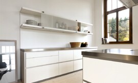 Deze moderne witte designkeuken maakt met zijn roestvrijstalen aanrechtbladen en greeploze witte fronten een strakke indruk. Aan de wand is een keukenblok geplaatst met grote, praktische lades. De lage kast daarboven heeft verschuifbare matglazen fronten en is voor een deel open. Het kookeiland heeft hetzelfde design: roestvrijstalen werkblad, witte greeploze fronten. Niets verstoort hier de kalmte die deze keuken uitstraalt. Zuordnung: Stil Design-keukens, Planungsart U-vormige keuken