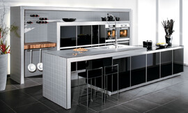 Moderne designkeuken met licht grijs betegeld aanrechtblad en wangen. De kasten zijn voorzien van hoogglans zwarte fronten. Het aanrechtblad is als het ware op de kasten gelegd en is daardoor iets hoger. Bovendien is er afstand bewaard ten opzichte van de kasten, zodat er een fraaie zitgelegenheid is ontstaan.

Het wandelement is betegeld met dezelfde tegels als die op het kookeiland gebruikt zijn. De kasten die in het wandelement opgenomen zijn hebben ook hier dezelfde zwarte hoogglans fronten. De apparatuur is op praktische werkhoogte naast elkaar aangebracht. Als extra luxe-element dient het butchter-block te worden genoemd, aangebracht naast de kasten in het wandelement. Daarboven bevindt zich de mogelijkheid de wijnverzameling op rekken te bewaren. Zuordnung: Stil Design-keukens, Planungsart L-vormige keuken