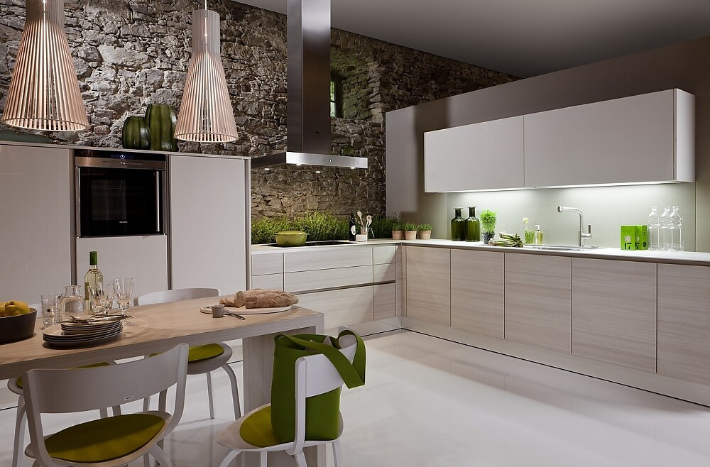 Deze lichtvoetige keuken in de L-vorm speelt met een subtiel contrast tussen lichte houttinten en gebroken witte vlakken. Fronten met greeplijsten houden de keuken optisch kalm. Er ontstaat een bijzonder contrast tussen de kalme design keuken en de ruwe stenen muur. Links zijn half hoge kasten geplaatst met daarin de apparatuur. Daarnaast is het fornuis met daaronder handige grote lades voor alle potten, pannen en ander kookgerei. Na de hoek komt er een blok met onderkasten, de spoelbak en daarboven de kasten met zeer brede deurtjes. Onder de hangkasten zijn handige spots aangebracht, zodat er altijd genoeg licht op het aanrechtblad schijnt. Zuordnung: Stil Klassieke keukens, Planungsart L-vormige keuken