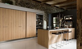U-vormige keuken met keukeneiland en zitgelegenheid. Optisch wordt de keuken gedragen door het ruwe eiken fineer met verticale tekening. Een robuust oppervlakte contrasteert mooi met de koele rvs-apparatuur. Het keukeneiland is van een dun zwart glanzend aanrechtblad voorzien. Zuordnung: Stil Klassieke keukens, Planungsart Keuken met keukeneiland