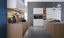 Deze keuken die uit drie elementen bestaat biedt een  combinatie van bijzonder natuurlijk werkende kopereiken fronten met matte sahara-lak oppervlakken. Het eerste keukenblok bevat het fornuis en de spoelbak. De kopereiken onderkasten zijn zwevend aangebracht. Daarboven hangen de bovenkasten in een witte kleur. In de hangkast is een efficiëntie afzuigkap geïntegreerd die geen luchtafvoer behoeft. Perfecte plaats om te werken in de keuken biedt het keukeneiland. Dit eiland is ook van kopereiken en staat op drie open metalen poten. Het andere wandblok, in dezelfde lichte kleur als de hangkasten, is hoog en biedt behalve veel bergruimte ook de nodige apparatuur. Midden in het hoge keukenblok bevindt zich een open kast, waar weer de kopereiken tint in terugkomt. Al met al een moderne keuken met een harmonische en rustige uitstraling met voldoende berg- en werkruimte. Zuordnung: Stil Moderne keukens, Planungsart L-vormige keuken