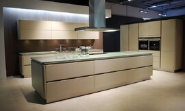Ruime eilandkeuken met kookveld op het kookeiland. Beige fronten met greeplijsten, afgewerkt met rondingen. Dun glazen aanrechtblad, kleurtint richting groen. Zuordnung: Stil Design-keukens, Planungsart Keuken met zitgelegenheid