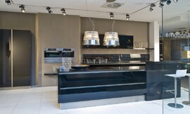 Deze moderne zwarte eilandkeuken in hoogglans uitvoering biedt behalve het kookeiland een keukenblok aan de wand met de spoelbak, onder- en bovenkasten. De bar aan het kookeiland biedt zitgelegenheid voor twee personen. De apparatuur is op praktische werkhoogte in de wand opgenomen. De koel- vriescombinatie is in separaat geplaatste hoge kasten geplaatst. De greeploze zwarte hoogglans fronten reflecteren licht en het patroon van de vloer. Hier ziet u ook een dubbele afzuigkap in een lampenkap. Zuordnung: Stil Moderne keukens, Planungsart keukenblok
