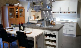 Moderne keuken met kookeiland en open rek voor kookgerei daarboven. Het kookeiland heeft een zithoek met stoelen op normale hoogte aangevoegd en een ingebouwd wijnrek. Zuordnung: Stil Moderne keukens, Planungsart Binneninrichting van de keuken