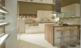 In deze U-vormige keuken is gebruik gemaakt van frisse witte fronten gecombineerd met een houten aanrechtblad dat aan de zijkant van het kookeiland doorloopt en ook als achtergrond bij een legplank is gebruikt. Aan de linkerkant zijn twee onderkasten opgenomen waarvan het bovenste gedeelte open is. Daarnaast komt de spoelkast en nog een onderkast. Boven de spoelbak zijn drie hangkastjes opgenomen. Links van de hangkastjes is een legplank bevestigd tegen hetzelfde hout als het aanrechtblad. Het andere wandgedeelte bestaat uit onderkasten, handige werkruimte op het aanrechtblad met daarboven twee brede kasten met glazen inzet en daaronder spotjes. Haaks hierop staat het kookeiland met fornuis, afzuigkap en aan de andere kant een gezellige zitgelegenheid. In de hoge wandkasten is de apparatuur opgenomen. Zuordnung: Stil Landelijke keukens, Planungsart U-vormige keuken