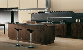 Design-eilandkeuken met moderne ronde vormen en dun werkblad. De keuken bestaat ten eerste uit een wandblok met onderkasten en fornuis. De spatwand bestaat uit hetzelfde zwarte materiaal als het aanrechtblad. De hoogte van het aanrecht varieert op speelse wijze. Daar tegenover is het keukeneiland geplaatst met de spoelbak. Het aanrechtblad loopt aan de andere kant iets op en vormt daardoor een bar met zitgelegenheid op barkrukken. De keuken biedt een interessant contrast tussen mat ebbenhout met verticaal patroon en zwarte designelementen. Verlichting met verschillende spots die geplaatst zijn in een hangend gedeelte boven het keukeneiland en in een doorlopende rand die in het wandgedeelte is opgenomen. Zuordnung: Stil Design-keukens, Planungsart Keuken met keukeneiland