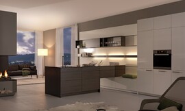 Een keuken die er niet meer direct als keuken uitziet, waardoor de keuken als deel van de woonruimte geplaatst kan worden. Een open en flexibel wandpaneel-kastsysteem vormt hier het middelpunt van de woonervaring en is tegelijkertijd deel van de keuken. De keuken bestaat ten eerste uit het witte wandpaneel waar kastjes aan opgehangen kunnen worden. Daarnaast zijn hoge witte kasten met de apparatuur geplaatst. Het kookeiland ten slotte is ook tegen het wandpaneel geplaatst en leunt op lage onderkasten, waarop een kleine zitgelegenheid mogelijk is geworden. Zuordnung: Stil Klassieke keukens, Planungsart Keuken met keukeneiland
